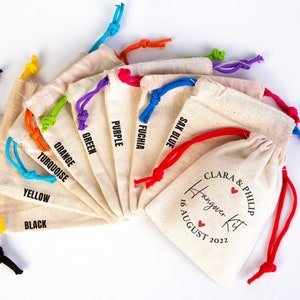 May include: A set of ten small, white cotton drawstring bags with colorful drawstrings. Each bag has a different color drawstring and a black text label with the color name. The last bag has a red drawstring and a black text label that says "Clara & Philip Hangover Kit 16 August 2022".