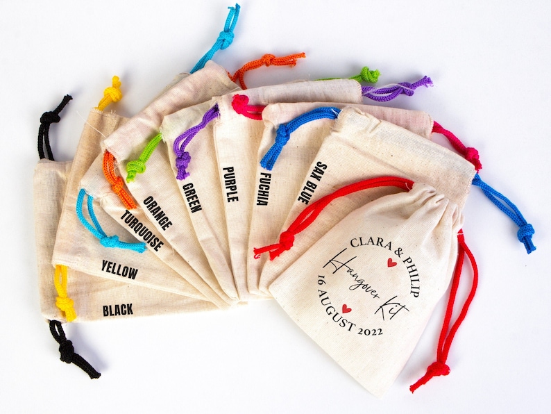 May include: A set of ten small, white cotton drawstring bags with colorful drawstrings. Each bag has a different color drawstring and a black text label with the color name. The last bag has a red drawstring and a black text label that says "Clara & Philip Hangover Kit 16 August 2022".