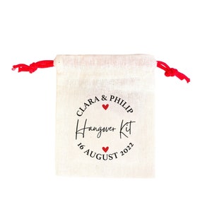 May include: A white cotton drawstring bag with a red drawstring. The bag has a black circle with the text "Clara & Philip" and a red heart. The text "Hangover Kit" is written in cursive below the circle. The date "16 August 2022" is written below the text.