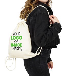 Peut inclure: Un sac à cordon blanc avec le texte "YOUR LOGO OR IMAGE HERE" en lettres vertes. Le sac est porté par une personne aux cheveux longs bruns et une veste en jean noire.