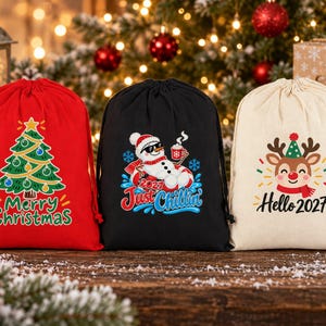 May include: Three drawstring gift bags in red, black, and cream. The red bag features a green Christmas tree and the text Merry Christmas. The black bag shows a snowman with sunglasses and the text 2021 Just Chillin. The cream bag displays a reindeer and the text Hello 2027.