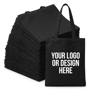 Pode incluir: Uma grande pilha de sacolas de algodão preto com duas alças. Uma sacola está posicionada na frente, apresentando o texto impresso em branco YOUR LOGO OR DESIGN HERE em uma fonte negrito sem serifa, centralizado na frente do tecido.
