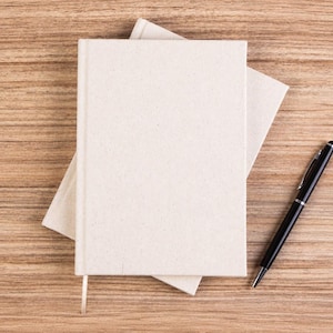 May include: Two stacked rectangular notebooks with plain, light beige fabric covers and a single thin ribbon bookmark extending from the bottom, accompanied by a sleek black ballpoint pen with a silver clip positioned to the right side.