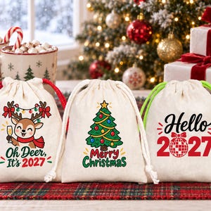 Può includere: Tre sacchetti in tela color avorio con coulisse e grafiche natalizie. Il sacchetto a sinistra mostra una renna con il testo "Oh Deer, It's 2027". Il sacchetto centrale mostra un albero di Natale con "Merry Christmas". Quello a destra dice "Hello 2027".