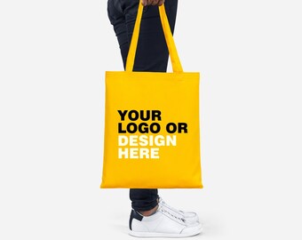 Può includere: Una persona tiene in mano una borsa di tela gialla con manici neri. La borsa ha la scritta "YOUR LOGO OR DESIGN HERE" stampata su di essa.