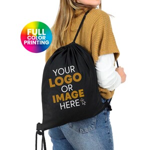 Op de afbeelding: Een zwarte trekkoordtas met de tekst "YOUR LOGO OR IMAGE HERE" in gouden letters. De tas wordt door een persoon gedragen. De tas is beschikbaar voor full colour bedrukking.