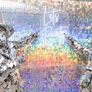 May include: Two people dressed in silver mirror costumes stand in front of a shimmering iridescent wall. The wall is covered in small square tiles. The people are pointing at the wall, which has the words "Let's Party" written in white neon letters.