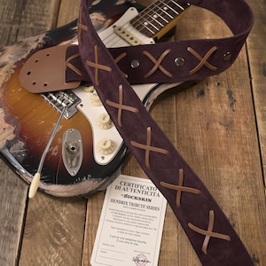 Könnte beinhalten: Ein dunkelvioletter Gitarrengurt mit braunen Lederkreuzdetails. Der Gurt liegt über einer Gitarre mit weißem Schlagbrett. Ein Echtheitszertifikat ist ebenfalls sichtbar.