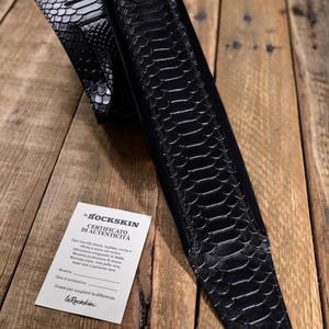 May include: Black guitar strap with a textured, snake-like pattern. The strap is made of leather and has a glossy finish. A white card with Italian text is visible. The strap is lying on a wooden surface.