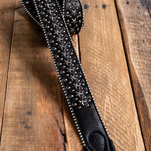 Könnte beinhalten: Ein schwarzer Gitarrengurt aus Leder mit einer mittleren Reihe aus silberfarbenen Metallsternen, flankiert von zwei Reihen kleiner, runder silberner Nieten. Die Kanten sind mit winzigen silbernen Perlen besetzt und das Ende ist verstärkt.