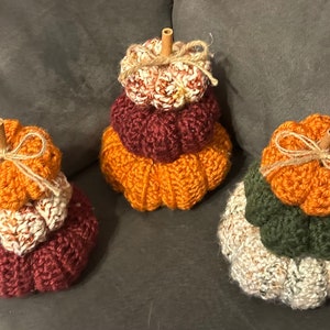 May include: Three crocheted pumpkins stacked on top of each other. The pumpkins are orange, burgundy, and white with a brown stem on top. The pumpkins are tied with twine.