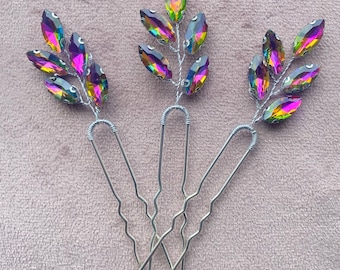 Puede incluir: Tres pinzas para el pelo iridiscentes con un diseño de hoja. Cada pinza tiene un alambre plateado con múltiples cristales en forma de lágrima en tonos de rosa, morado y verde.