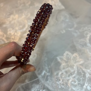 Könnte beinhalten: Ein burgunderfarbenes Perlenhaarband mit einer goldenen Drahtbasis. Das Haarband besteht aus kleinen, facettierten Perlen, die in einer durchgehenden Reihe aneinandergereiht sind.