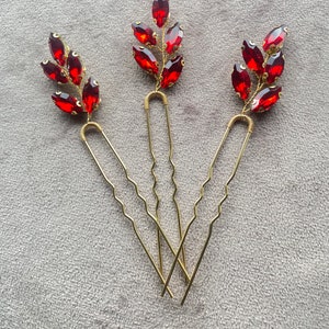 Può includere: Tre forcine dorate con motivi a foglia di pietre preziose rosse. Le forcine sono ondulate e hanno un anello decorativo in cima.