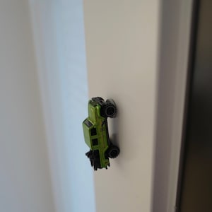 May include: A green toy car with black wheels is stuck to a white wall.