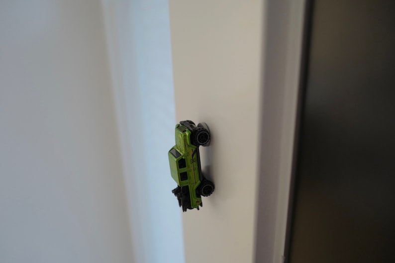 May include: A green toy car with black wheels is stuck to a white wall.