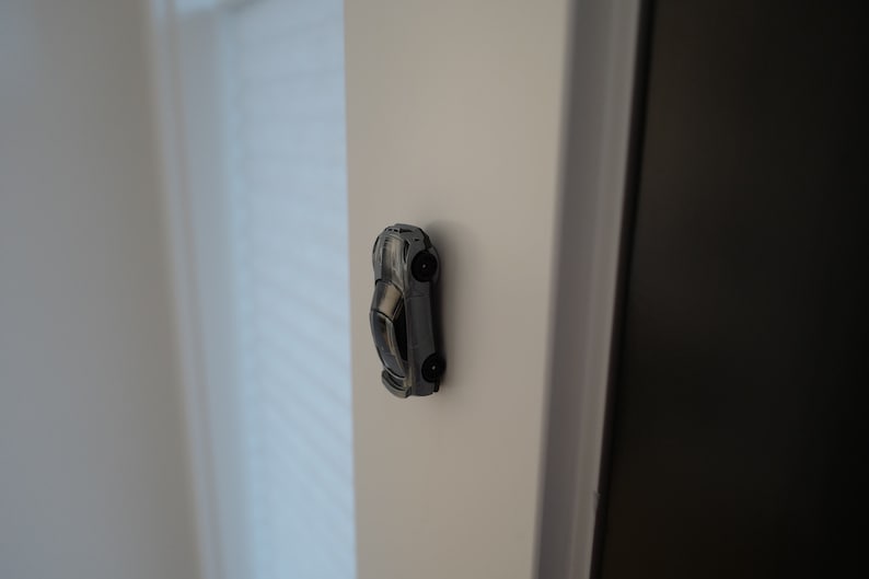 May include: A silver toy car is attached to a white wall with a magnet.