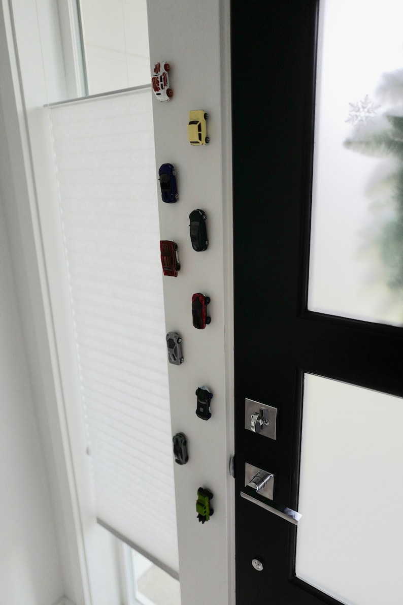 May include: A white door with a black door frame and a row of toy cars attached to the door with magnets. The cars are in various colors, including red, blue, yellow, and green.