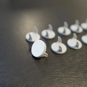 May include: A group of nine white plastic hooks with a round base and a curved hook. The hooks are arranged in a line on a dark gray textured surface.