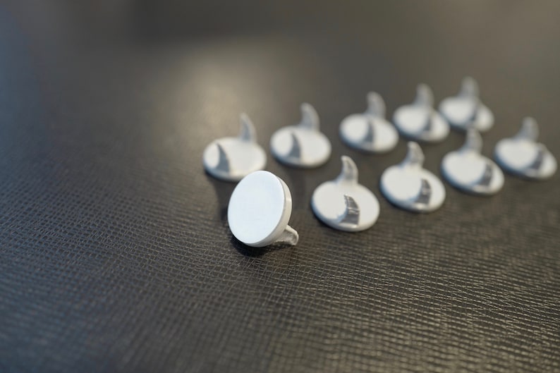 May include: A group of nine white plastic hooks with a round base and a curved hook. The hooks are arranged in a line on a dark gray textured surface.