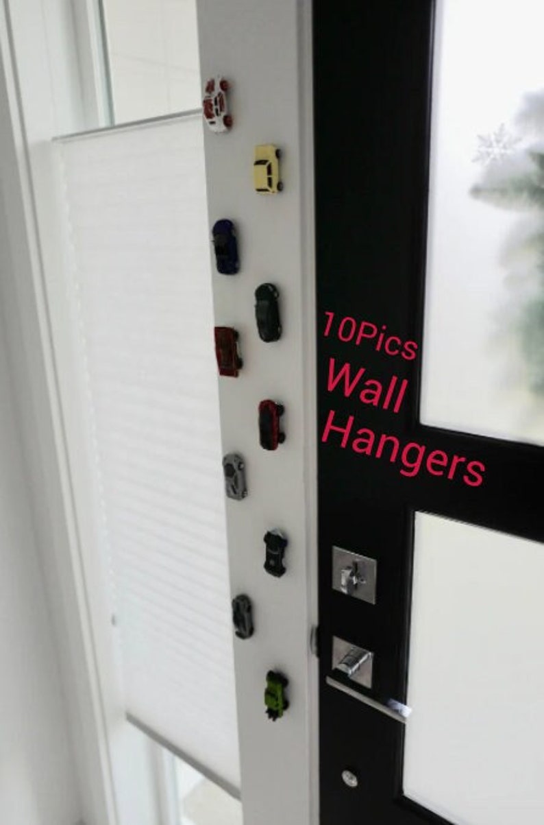 May include: A white wall with a row of small toy cars attached to it with magnets. The cars are in different colors, including red, blue, yellow, and black. The text "10Pics Wall Hangers" is written on a black door behind the cars.