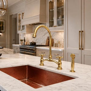 May include: A rectangular hammered copper kitchen sink set into a white marble worktop. The sink features a high-arc brass tap with two cross-handle controls and a matching side spray, creating a functional and metallic focal point.