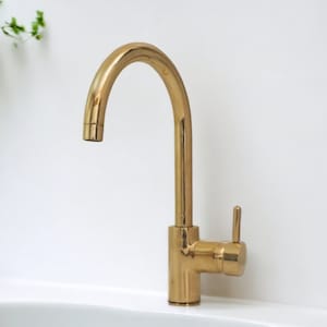May include: A gold-toned kitchen faucet with a single lever handle and a curved spout. The faucet is mounted on a white countertop.