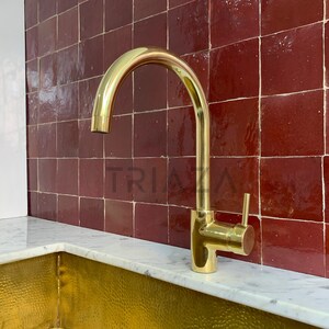 May include: A gold-toned kitchen faucet with a single handle and a high arc spout. The faucet is mounted on a white countertop in front of a red tile backsplash.