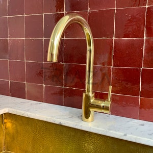 May include: A gold-toned kitchen faucet with a single handle and a high arc spout. The faucet is mounted on a white countertop in front of a red tile backsplash.
