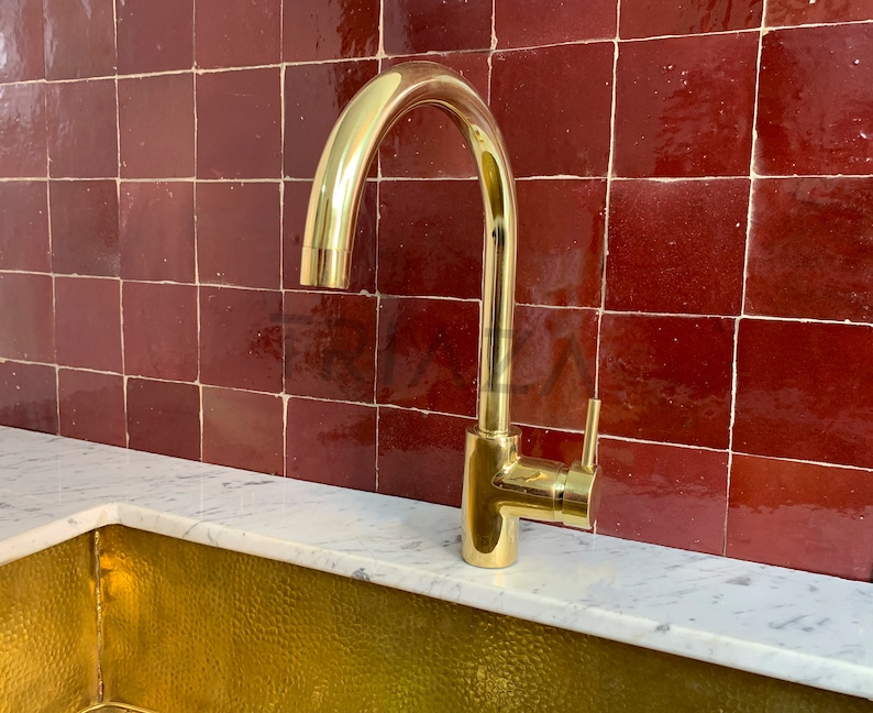 May include: A gold-toned kitchen faucet with a single handle and a high arc spout. The faucet is mounted on a white countertop in front of a red tile backsplash.