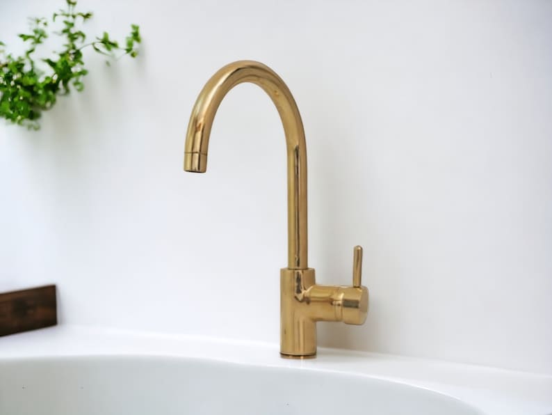 May include: A gold-toned kitchen faucet with a single lever handle and a curved spout. The faucet is mounted on a white countertop.