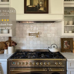 May include: A white kitchen with white cabinets and a white tiled backsplash. A stainless steel stove with gold accents is in the center of the image. A white teapot sits on the stovetop. The cabinets have glass doors and are filled with dishes and other kitchen items. The backsplash is made of small, square tiles. The stove has a digital display and multiple knobs. The countertop is white and has a marble-like pattern. The image includes the text "ONE OF TRIAZA'S PROJECTS" and "Happy client from California USA".