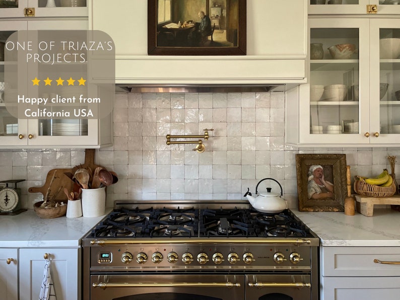 May include: A white kitchen with white cabinets and a white tiled backsplash. A stainless steel stove with gold accents is in the center of the image. A white teapot sits on the stovetop. The cabinets have glass doors and are filled with dishes and other kitchen items. The backsplash is made of small, square tiles. The stove has a digital display and multiple knobs. The countertop is white and has a marble-like pattern. The image includes the text "ONE OF TRIAZA'S PROJECTS" and "Happy client from California USA".