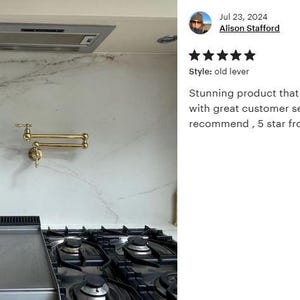 May include: A gold-toned kitchen faucet with a pull-down sprayer mounted on a white marble backsplash. The faucet has a lever handle and a swivel spout. The backsplash is accented with a gray veined pattern.  The faucet is installed above a black gas stovetop with a stainless steel griddle.