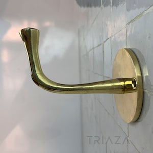 May include: A brass wall hook with a curved arm and a round base. The hook is mounted on a white tiled wall.