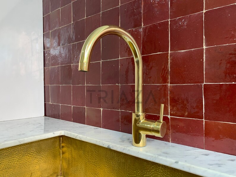 May include: A gold-toned kitchen faucet with a single handle and a high arc spout. The faucet is mounted on a white countertop in front of a red tile backsplash.