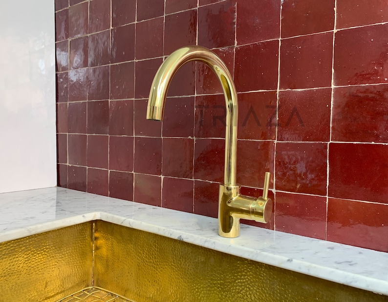 May include: A gold-toned kitchen faucet with a curved spout and a lever handle. The faucet is mounted on a white countertop in front of a red tile backsplash.