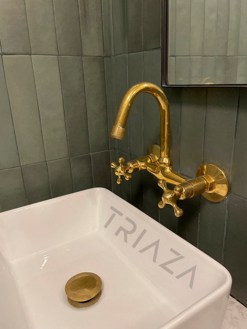 Puede incluir: Un lavabo de ba&ntilde;o de porcelana blanca con un tap&oacute;n de desag&uuml;e dorado. Un grifo dorado con manijas en forma de cruz est&aacute; montado en una pared de azulejos verdes detr&aacute;s del lavabo. El lavabo tiene la palabra "TRIAZA" impresa en el lateral.