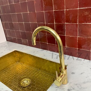 May include: A gold kitchen sink with a brass faucet against a backdrop of red tile.