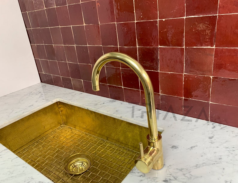 May include: A gold kitchen sink with a brass faucet against a backdrop of red tile.