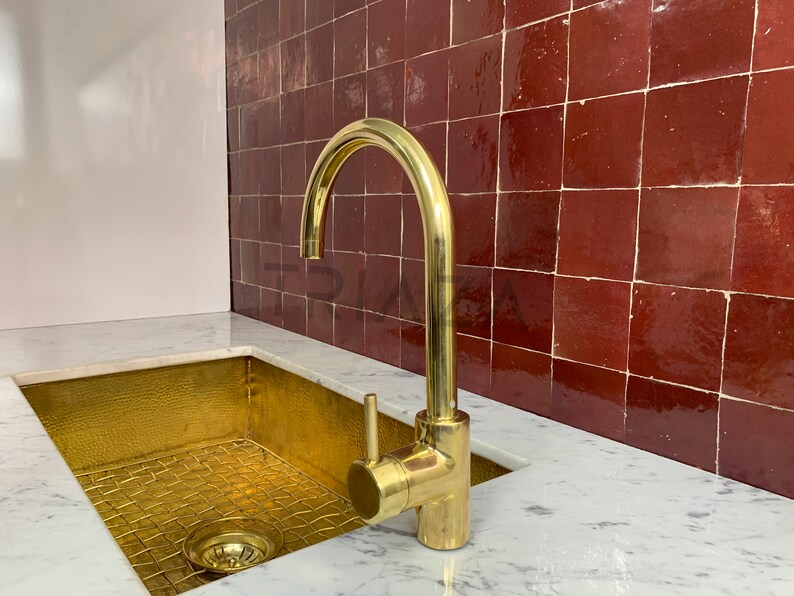 May include: A gold kitchen sink with a matching gold faucet against a backdrop of red tile.