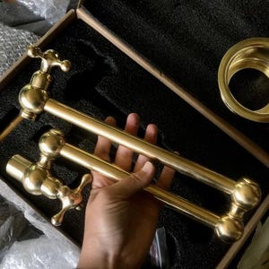 May include: Two gold-toned metal faucet spouts with round handles and a cross-shaped handle. The spouts are held in a hand.