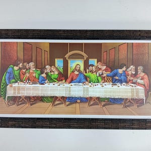 Op de afbeelding: Een ingelijst schilderij dat het Laatste Avondmaal voorstelt, een beroemde bijbelse scène. Het schilderij toont Jezus Christus en zijn twaalf discipelen die rond een tafel zitten, met een gedetailleerde achtergrond en een houten lijst.