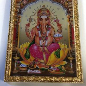 May include: A framed image of the Hindu deity Ganesha, depicted with a large head, elephant trunk, and four arms. He is seated on a lotus flower and holding a variety of objects, including a sweet, a book, and a bowl of sweets. The image is surrounded by a gold frame with intricate details.