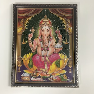 May include: A framed image of a Hindu deity, Ganesha, with a colorful background and a gold frame. The image is decorated with glitter and shows Ganesha sitting on a lotus flower.