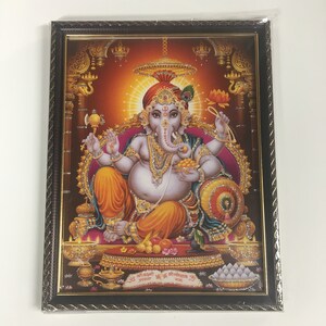 May include: Framed print of the Hindu deity Ganesha, depicted with an elephant head and multiple arms, wearing a red turban and gold jewelry. The artwork features vibrant colors and intricate details, set against a warm, reddish-orange background. The frame is dark brown.