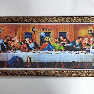 Op de afbeelding: Een ingelijst schilderij dat het Laatste Avondmaal voorstelt, een beroemde Bijbelse scène. Het schilderij toont Jezus Christus en zijn discipelen die rond een tafel zijn verzameld, met een houten achtergrond en een raam op de achtergrond.