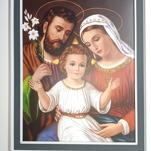 May include: A framed painting depicting the Holy Family.  Joseph, Mary, and Jesus are depicted in a traditional style with warm colors and soft features.  The painting is set against a dark background and includes a lily flower.