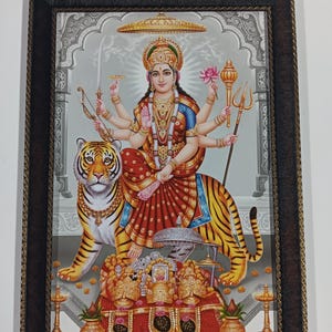 May include: A framed print depicting a multi-armed deity seated on a tiger. The deity wears a crown and holds various objects. The print features vibrant colors, including red, gold, and blue, and is set within a dark brown frame.