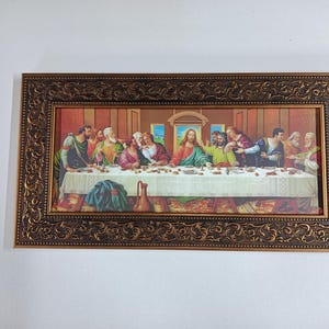 Op de afbeelding: Een ingelijst schilderij dat het Laatste Avondmaal voorstelt, een beroemde bijbelse scène. Het schilderij toont Jezus Christus en zijn discipelen die rond een tafel zijn verzameld, met een gedetailleerde achtergrond en ingewikkelde details.